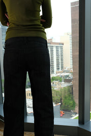 Person wearing a green top and black pants standing by a window with a cityscape view.