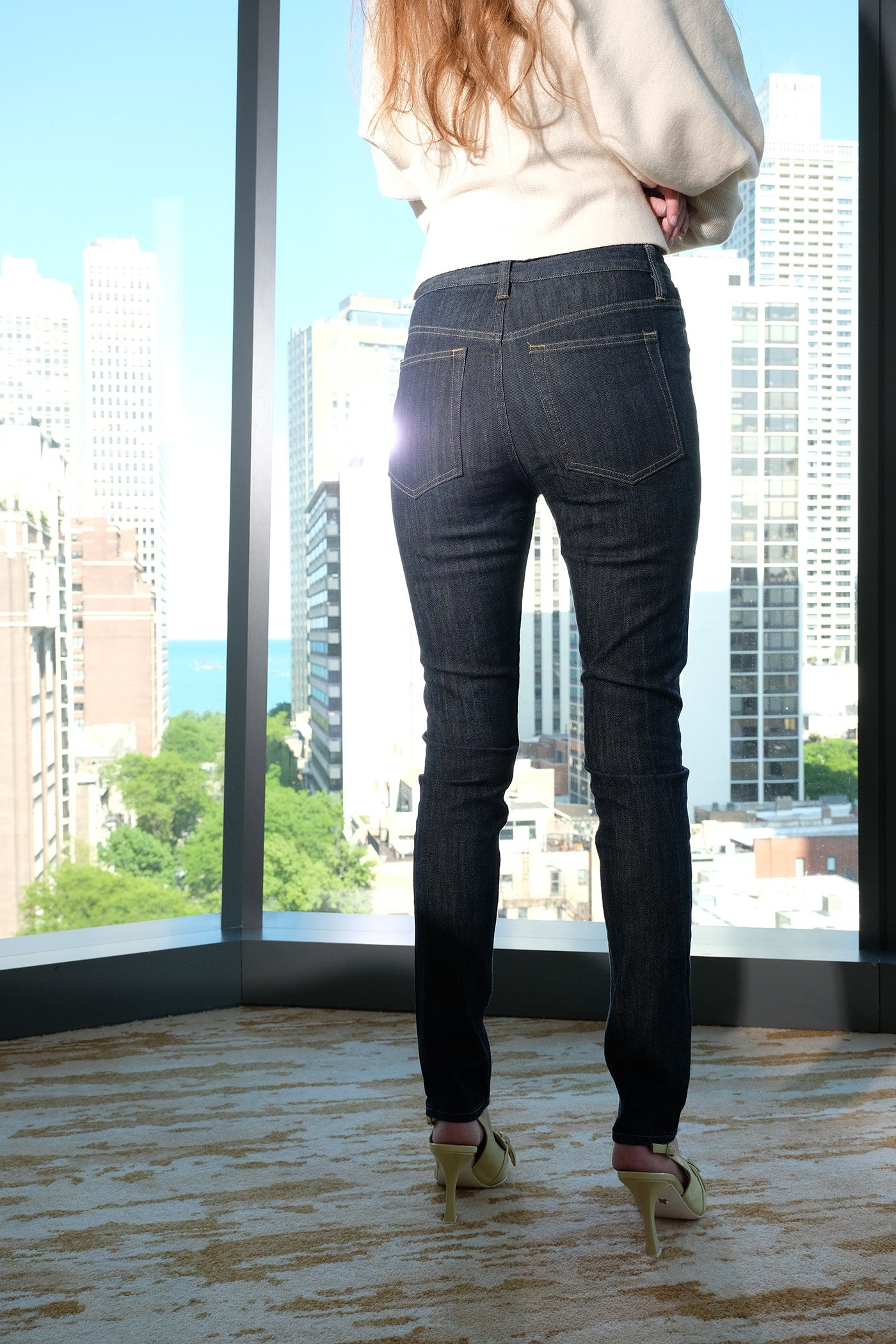 Person wearing dark jeans and high heels standing in front of a large window with cityscape view.