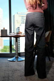 Person wearing dark blue jeans standing in a room with large windows showing cityscape.