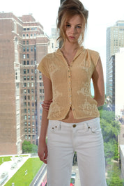 Woman wearing a beige knitted top and white pants standing in an urban setting with tall buildings.